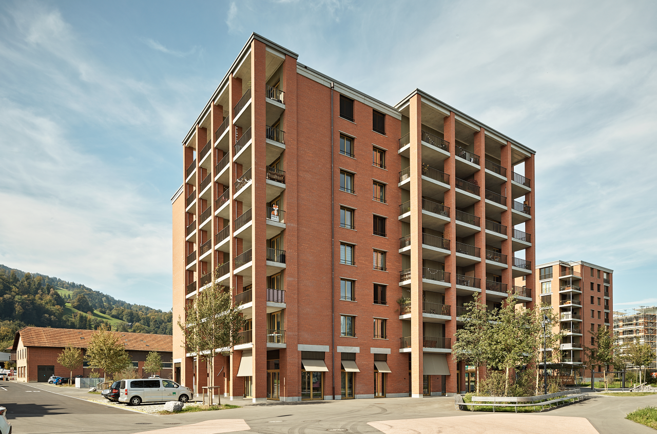 Rotes Mehrfamilienhaus mit Balkonen, davor parken Autos, im Hintergrund grüne Hügel.