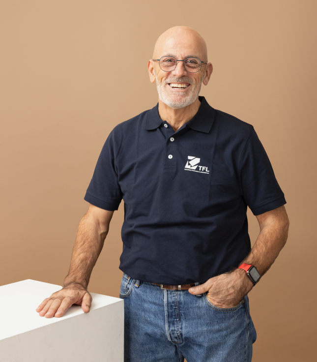 Aelterer Mann mit Brille und Bart laechelt, traegt dunkelblaues Polohemd und Jeans.